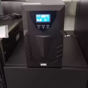 Front view of a black tower Uninterruptible Power Supply (UPS) sitting on a dark desk or shelf in a working environment, with its blue LCD screen illuminated and active.