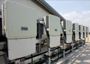 A row of large, grey solar power inverters mounted on the exterior wall of a building, used for converting direct current (DC) to alternating current (AC).