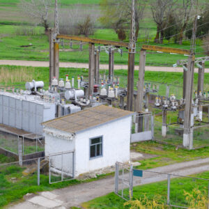 An outdoor electrical substation in a grassy rural setting, featuring high-voltage transformers, power line structures, and a small white control building.