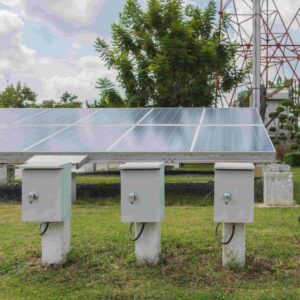 A ground-mounted solar panel installation in a grassy field, featuring three grey electrical control boxes mounted on short concrete pillars in the foreground.