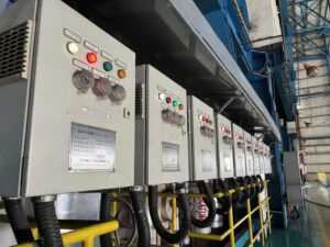 A line of grey industrial electrical control panels with buttons and indicator lights, mounted along a yellow railing inside a factory or industrial facility.