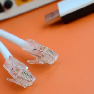A close-up of two white Ethernet cable ends with clear RJ45 connectors lying on a bright orange surface, with a blurred USB device and electronic equipment in the background.